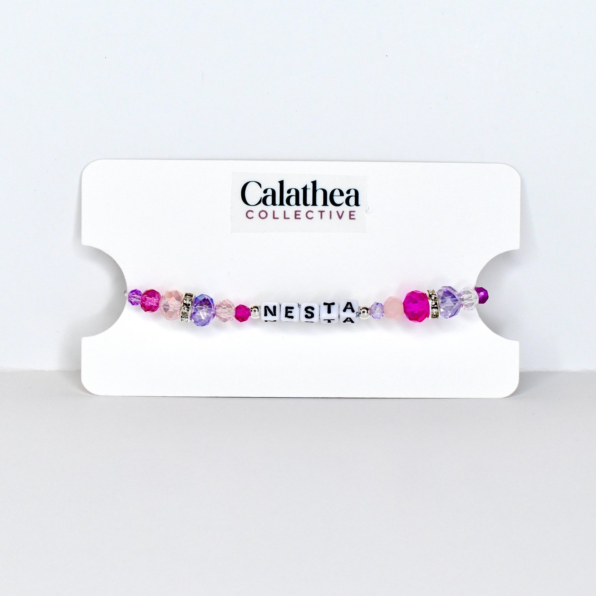 A beaded bracelet with the name "NESTA" spelled out in white letter beads. The bracelet features a mix of pink, purple, and clear beads, with a few silver accent beads. The bracelet is displayed on a white card with the "Calathea Collective" logo.