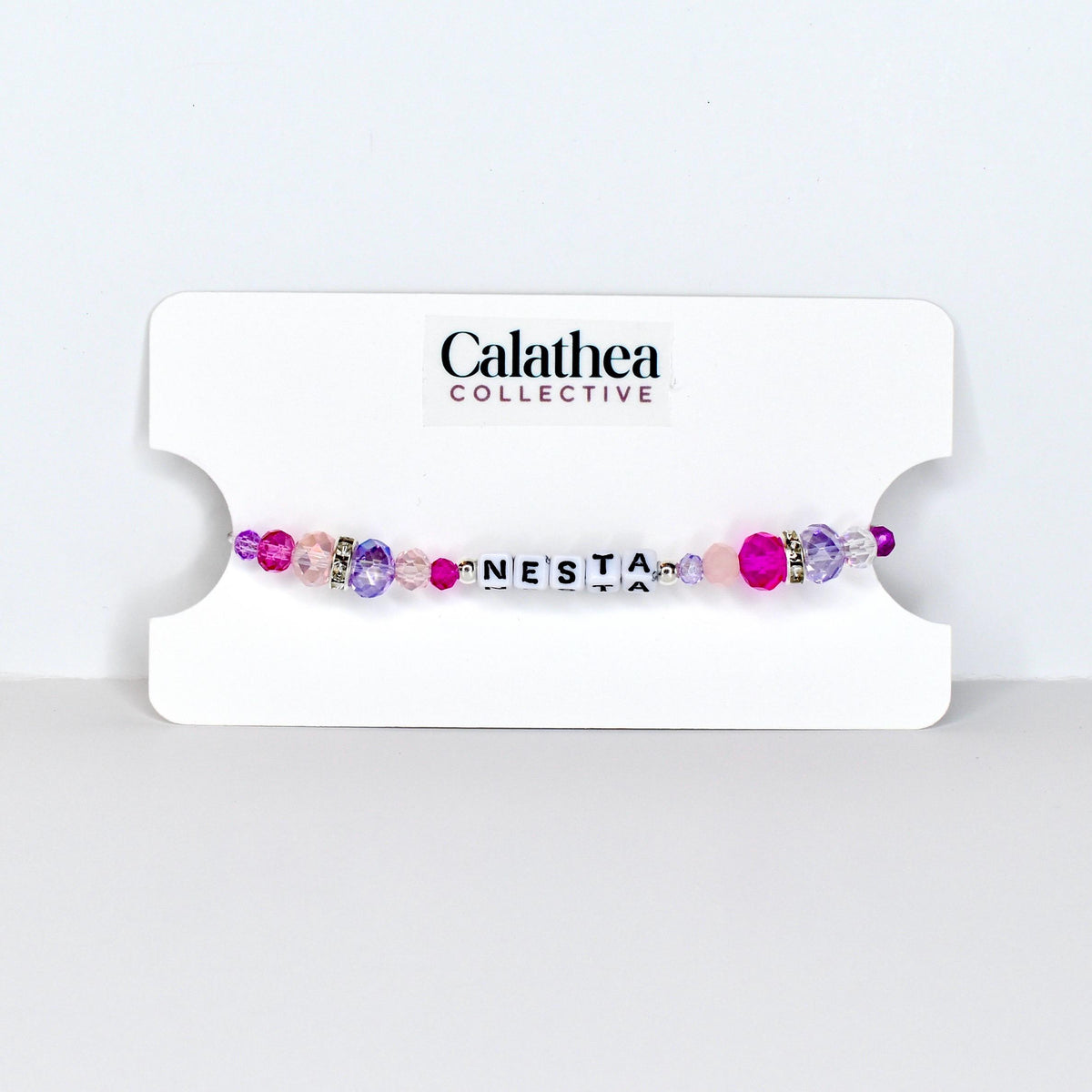 A beaded bracelet with the name "NESTA" spelled out in white letter beads. The bracelet features a mix of pink, purple, and clear beads, with a few silver accent beads. The bracelet is displayed on a white card with the "Calathea Collective" logo.