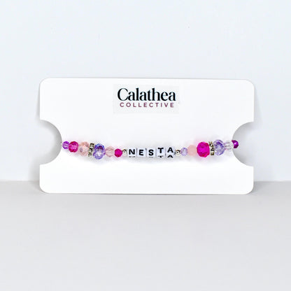 A beaded bracelet with the name "NESTA" spelled out in white letter beads. The bracelet features a mix of pink, purple, and clear beads, with a few silver accent beads. The bracelet is displayed on a white card with the "Calathea Collective" logo.