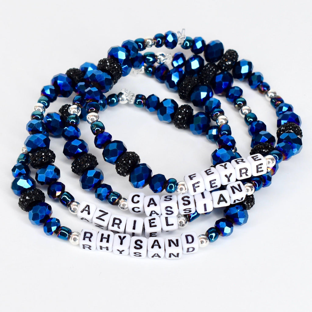 Four beaded bracelets with blue and black faceted beads and silver accents. Each bracelet has white letter beads spelling out names: Feyre, Cassian, Azriel, and Rhysand. The bracelets are on a white surface.