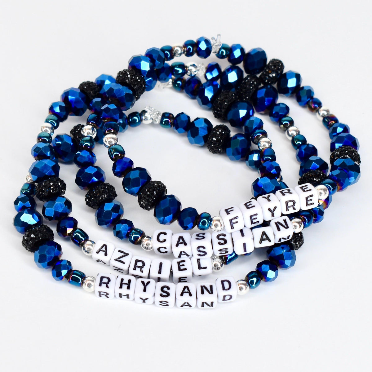 Four beaded bracelets with blue and black faceted beads and silver accents. Each bracelet has white letter beads spelling out names: Feyre, Cassian, Azriel, and Rhysand. The bracelets are on a white surface.