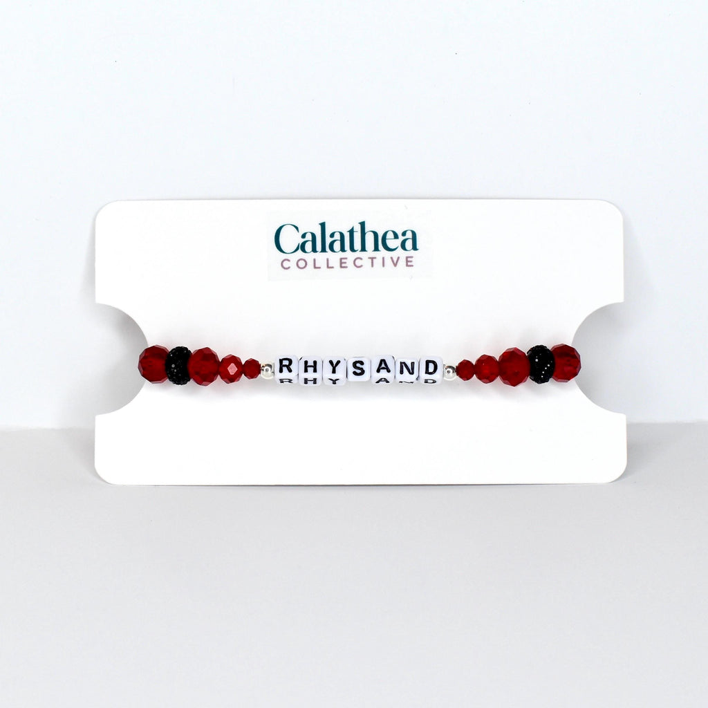 A beaded bracelet with the name "RHYSAND" spelled out in white letter beads. The bracelet features red and black beads, and is displayed on a white card with the "Calathea Collective" logo.