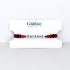 A beaded bracelet with the name "RHYSAND" spelled out in white letter beads. The bracelet features red and black beads, and is displayed on a white card with the "Calathea Collective" logo.