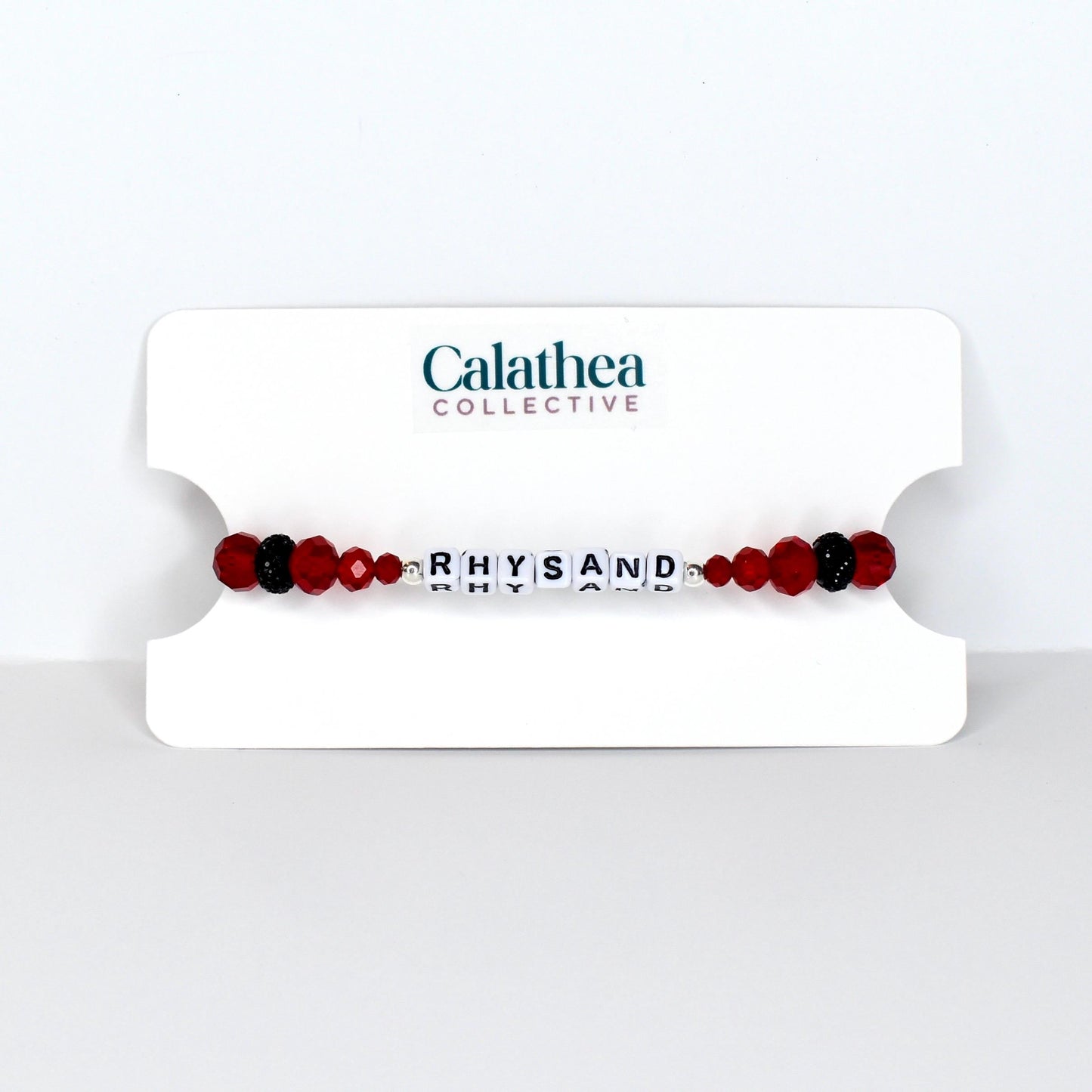 A beaded bracelet with the name "RHYSAND" spelled out in white letter beads. The bracelet features red and black beads, and is displayed on a white card with the "Calathea Collective" logo.