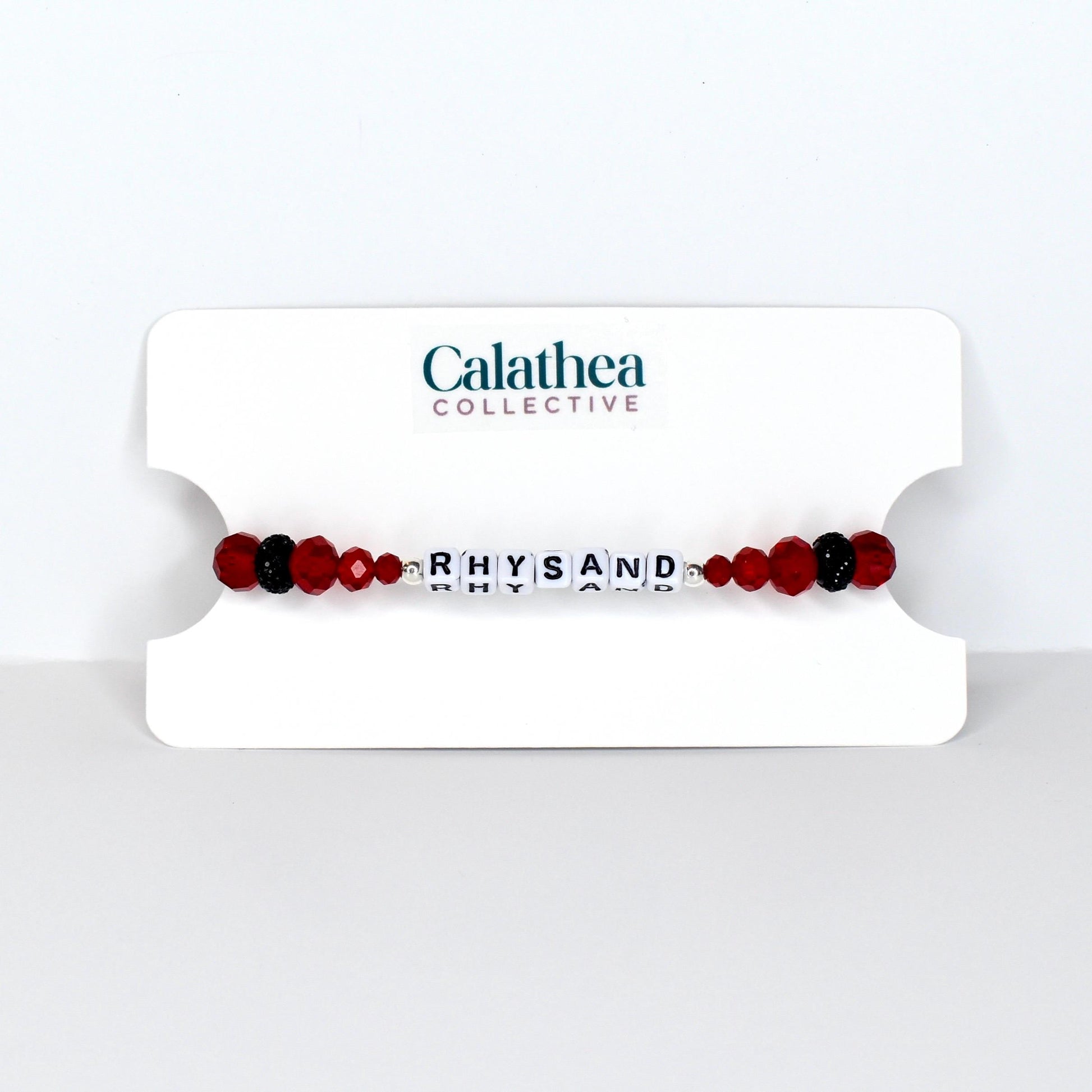 A beaded bracelet with the name "RHYSAND" spelled out in white letter beads. The bracelet features red and black beads, and is displayed on a white card with the "Calathea Collective" logo.