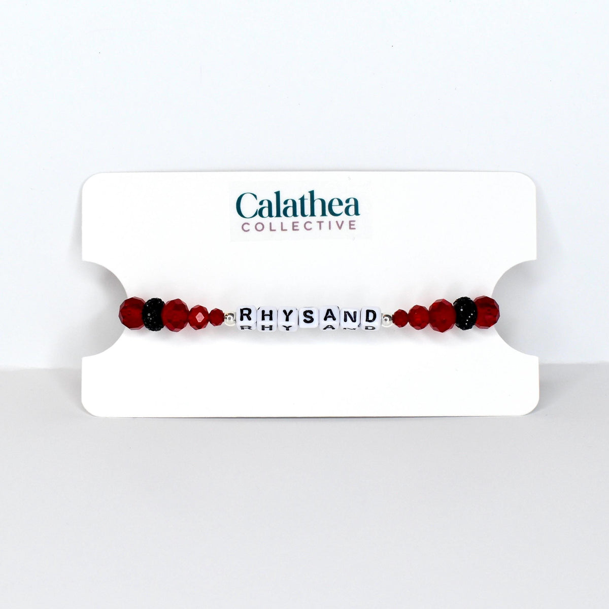 A beaded bracelet with the name "RHYSAND" spelled out in white letter beads. The bracelet features red and black beads, and is displayed on a white card with the "Calathea Collective" logo.