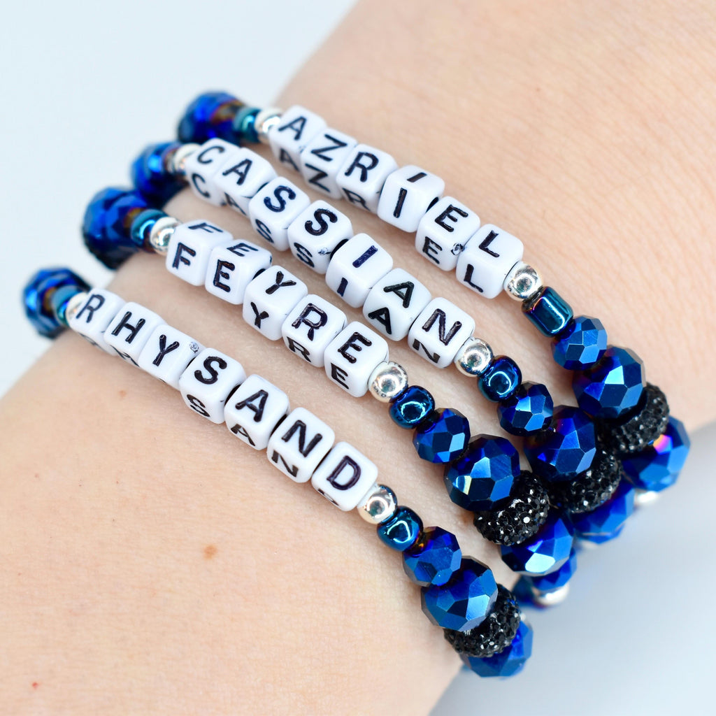 Four beaded bracelets with white lettered cubes spelling out names. The bracelets feature blue faceted beads, silver accents, and black rhinestone beads. The names include Azriel, Cassian, Feyre, and Rhysand.
