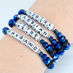 Four beaded bracelets with white lettered cubes spelling out names. The bracelets feature blue faceted beads, silver accents, and black rhinestone beads. The names include Azriel, Cassian, Feyre, and Rhysand.