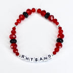A red beaded bracelet with the name "RHYSAND" spelled out in white letter beads. The bracelet features faceted red beads, interspersed with black, sparkly beads and silver accents. The bracelet is displayed on a white surface.