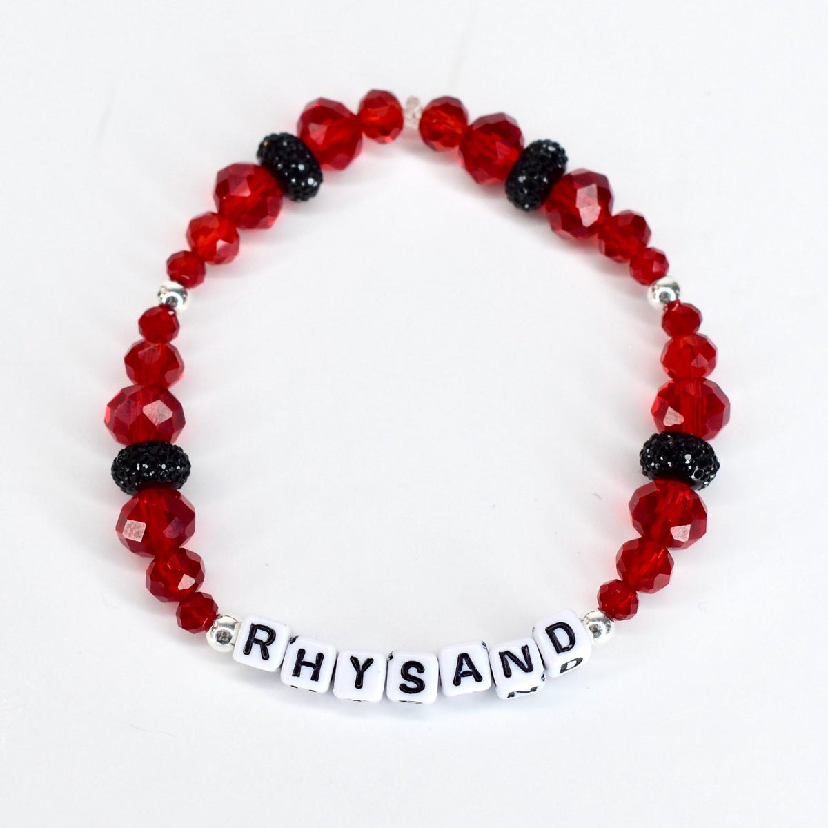 A red beaded bracelet with the name "RHYSAND" spelled out in white letter beads. The bracelet features faceted red beads, interspersed with black, sparkly beads and silver accents. The bracelet is displayed on a white surface.