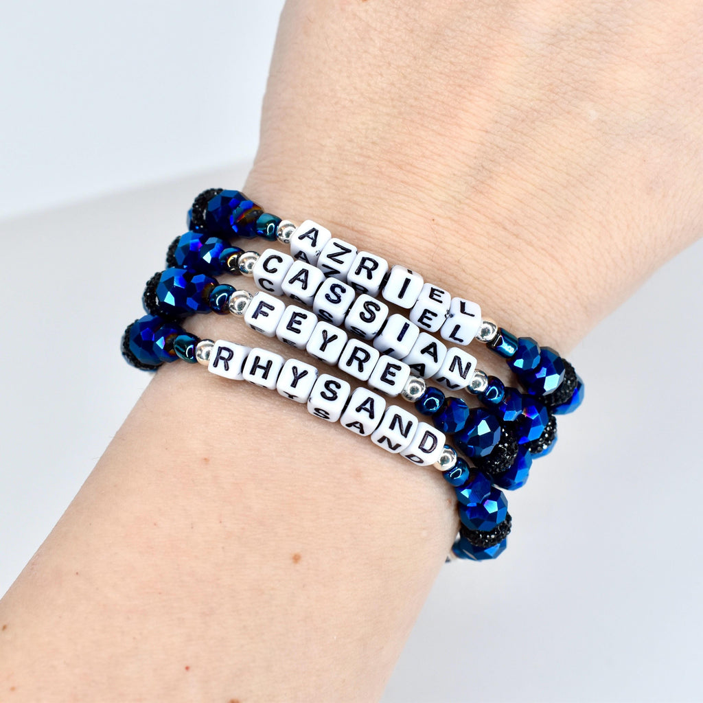 Four beaded bracelets with white lettered cubes spelling out names: Azriel, Cassian, Feyre, and Rhysand. The bracelets have dark blue faceted beads and black accents. The bracelets are worn on a wrist.