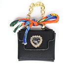 Black miniature handbag with a gold chain handle and a decorative scarf. The bag features a heart-shaped jewel with a gold frame. The scarf is orange, blue, and green.