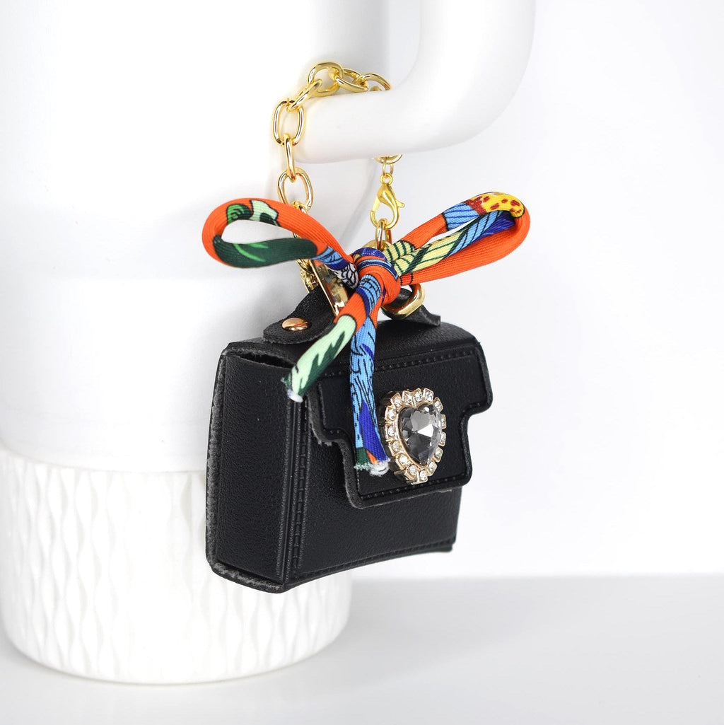A black handbag-shaped keychain with a gold chain and a colorful scarf tied to the top. The bag features a large, clear jewel with a gold border. The scarf has orange, green, blue, and yellow patterns.
