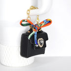 A black handbag-shaped keychain with a gold chain and a colorful scarf tied to the top. The bag features a large, clear jewel with a gold border. The scarf has orange, green, blue, and yellow patterns.