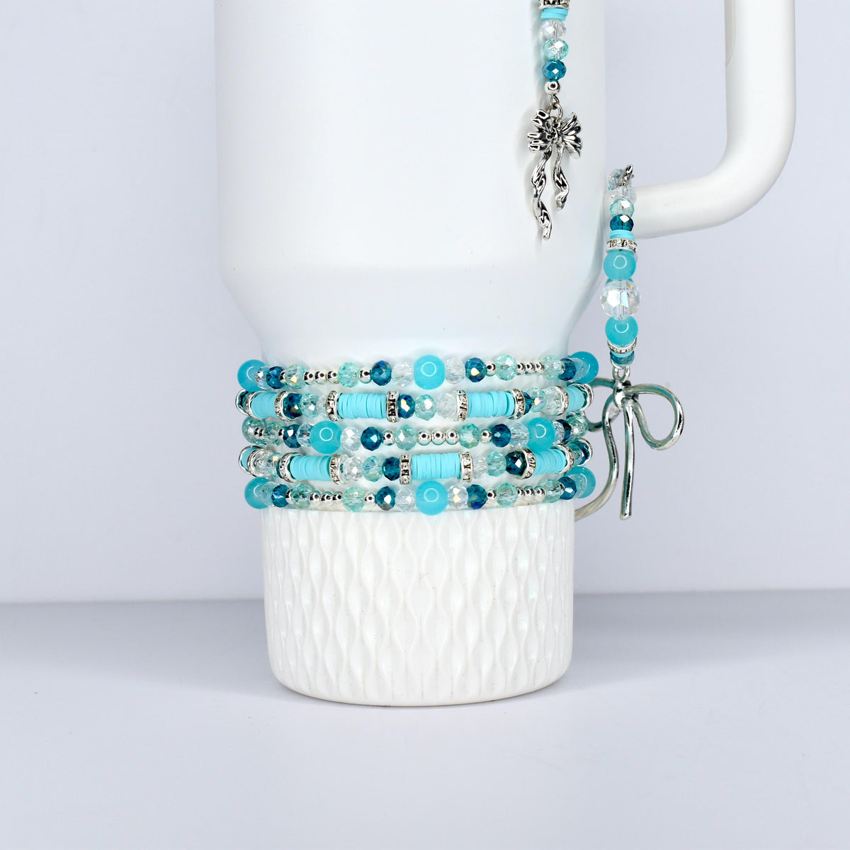 A white Stanley tumbler is adorned with multiple beaded bracelets in shades of turquoise, teal, and clear beads. A matching charm and bracelet hang from the handle, creating a cohesive jewelry set.