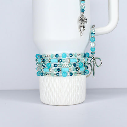 A white Stanley tumbler is adorned with multiple beaded bracelets in shades of turquoise, teal, and clear beads. A matching charm and bracelet hang from the handle, creating a cohesive jewelry set.