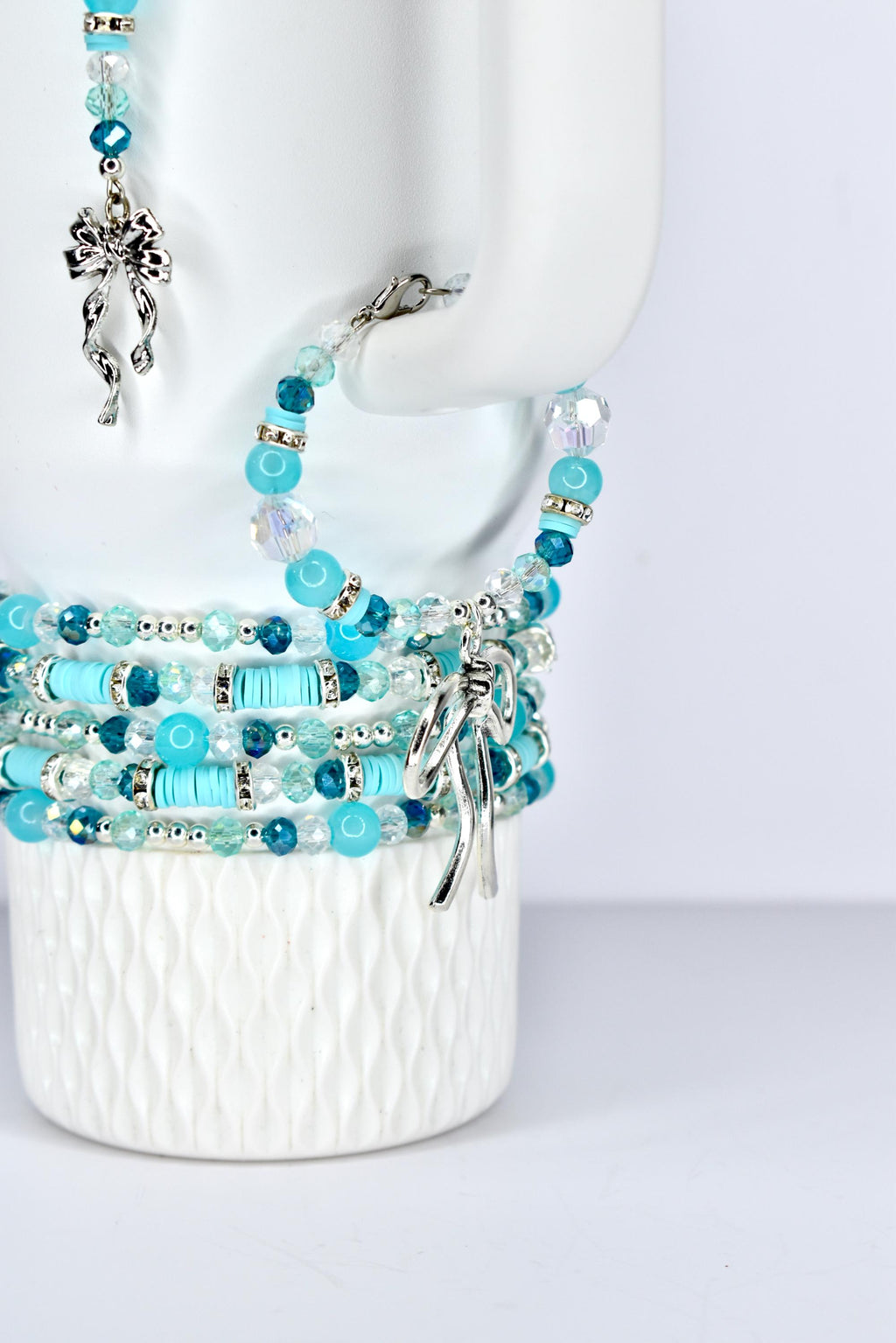 A collection of beaded bracelets and a tumbler handle charm in shades of blue and clear. The bracelets feature various bead sizes and shapes, with silver-colored bow charms. The jewelry is displayed on a white, textured surface.