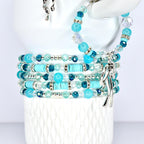 A stack of turquoise and blue beaded bracelets with silver accents. The bracelets are wrapped around a white cylindrical form. The beads include round, faceted, and cylindrical shapes. A silver charm hangs from one bracelet.