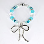 A beaded bracelet featuring a silver bow charm. The bracelet is composed of various shades of blue and clear beads, including round and faceted styles. The bow charm is a prominent, stylized design.