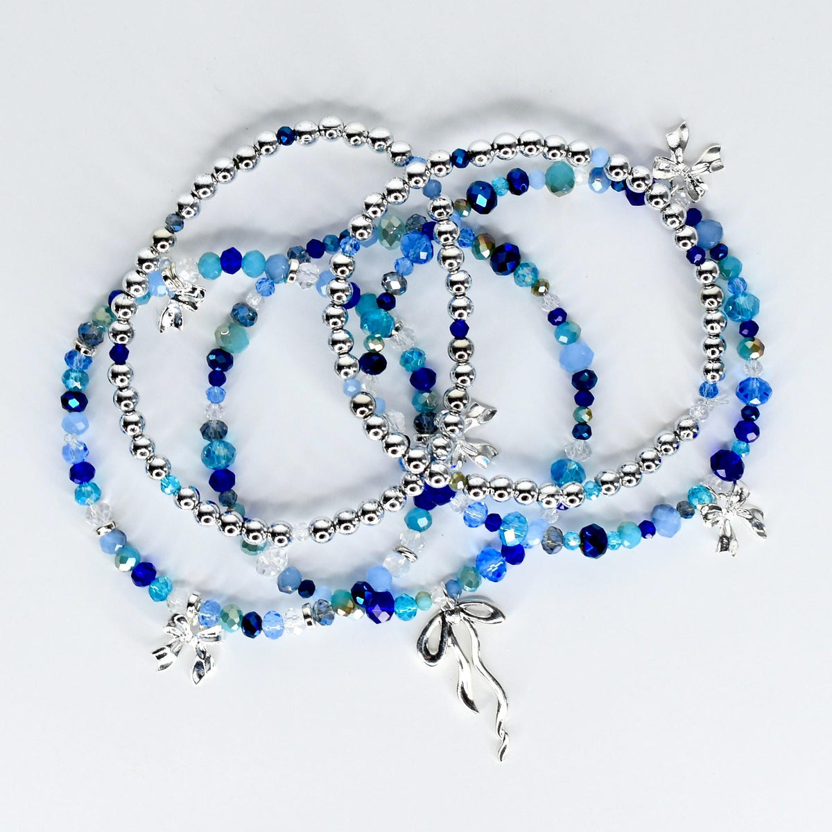 Several beaded bracelets in shades of blue and silver. The bracelets feature a mix of small, round silver beads and larger, faceted blue beads. Some bracelets have silver charms in the shape of flowers and a ribbon.