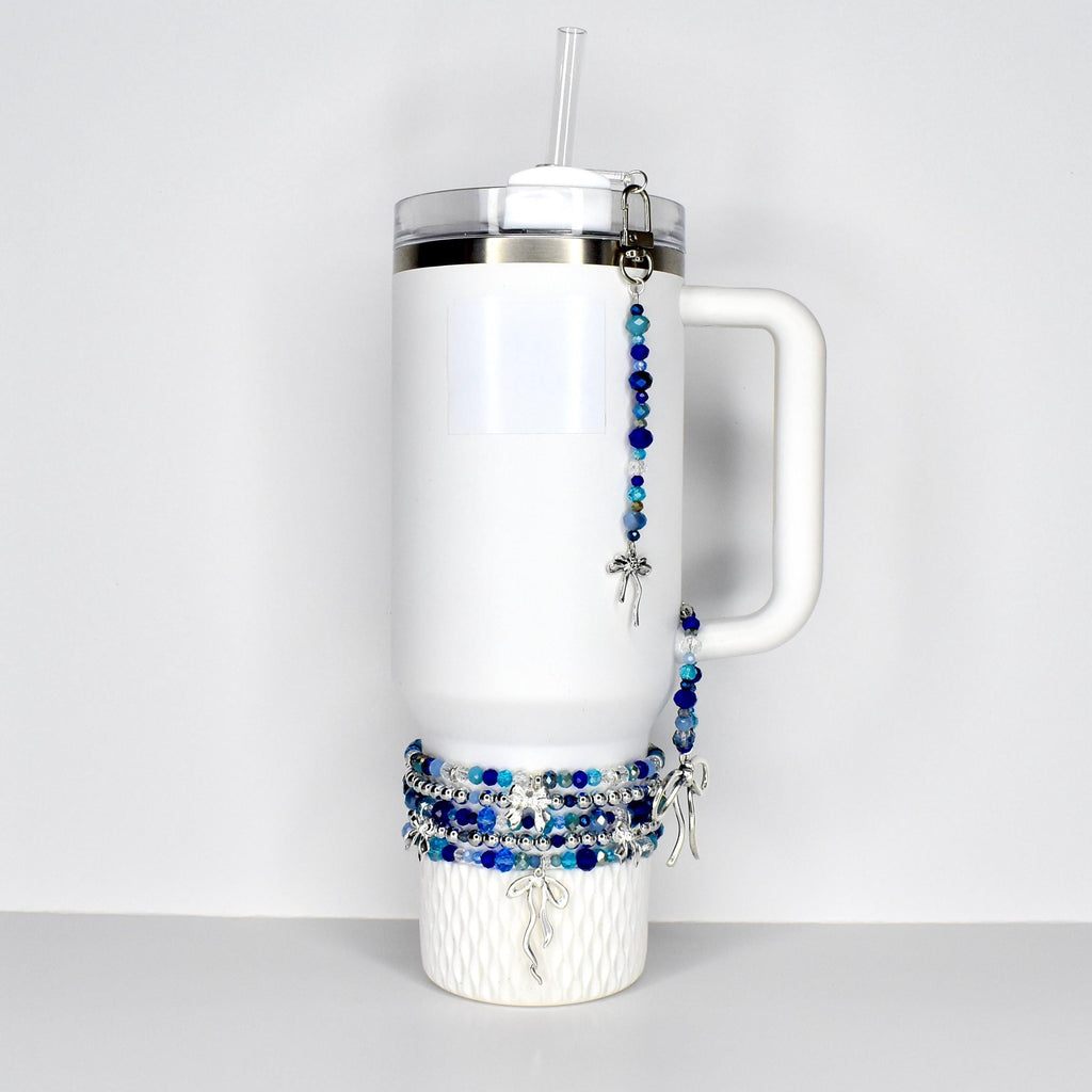 White insulated tumbler with a handle, a clear straw, and a silver-tone lid. The tumbler is adorned with beaded bracelets and a beaded charm with a silver-tone bow charm. The beads are shades of blue and clear.