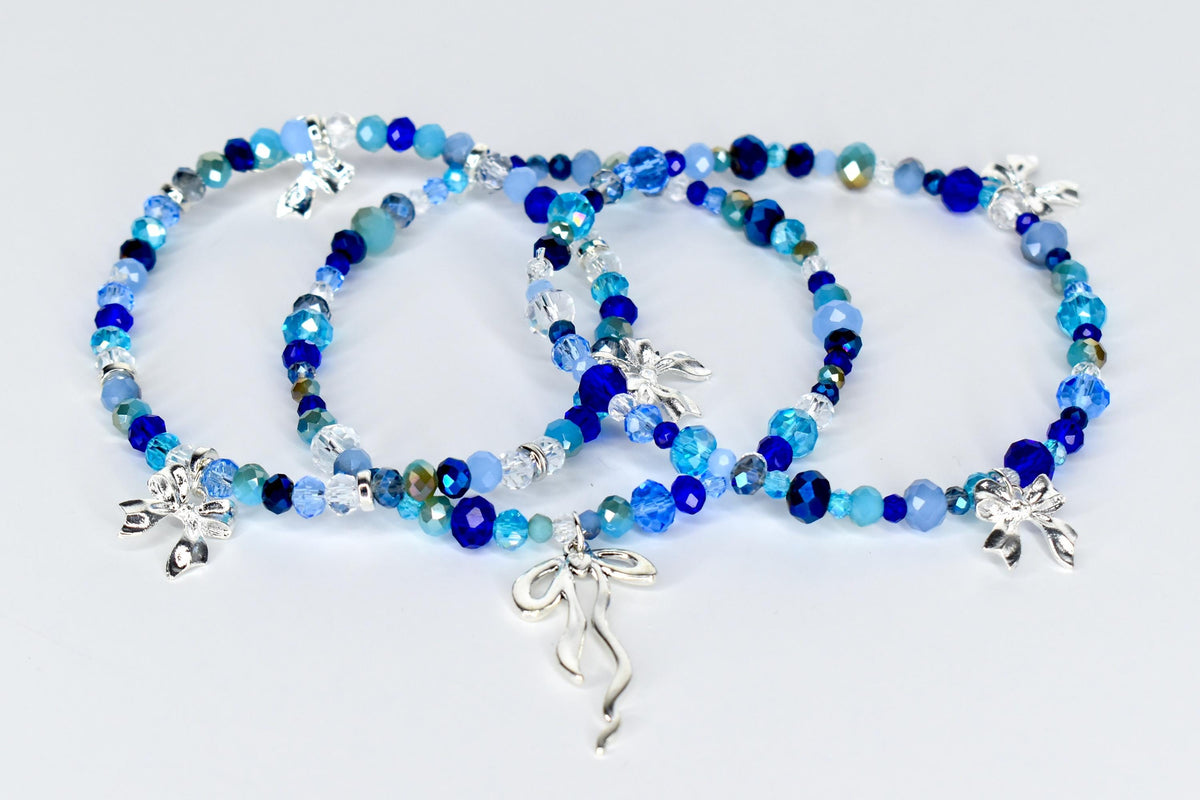 Three beaded bracelets in shades of blue, with silver bow charms. The bracelets feature a variety of blue beads, including faceted and round beads. The bracelets are displayed on a white surface.