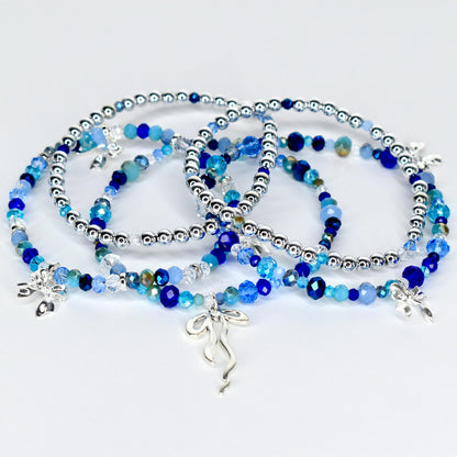 Several beaded bracelets in shades of blue and silver are displayed. The bracelets feature a mix of small, round silver beads and larger, faceted blue beads. Each bracelet has a silver charm. The bracelets are on a white surface.