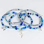 Several beaded bracelets in shades of blue and silver are displayed. The bracelets feature a mix of small, round silver beads and larger, faceted blue beads. Each bracelet has a silver charm. The bracelets are on a white surface.