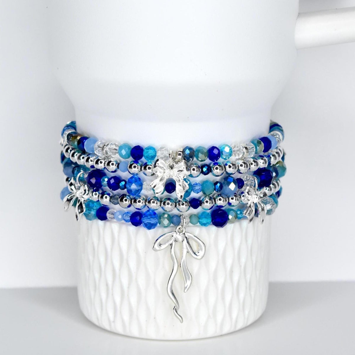 A stack of beaded bracelets in shades of blue and silver. The bracelets feature various sizes of beads and silver accents, including bow-shaped charms. The bracelets are displayed on a white Stanley tumbler.