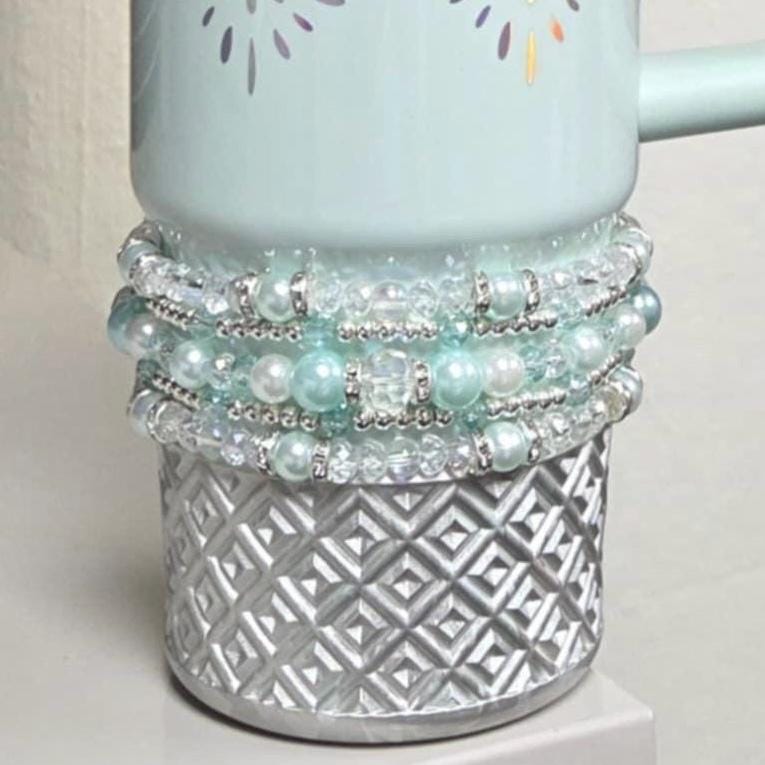 A light blue ceramic mug with a silver geometric patterned base. The mug is adorned with multiple strands of white and light blue beads and clear crystals. The top of the mug has a floral design.