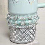 A light blue ceramic mug with a silver geometric patterned base. The mug is adorned with multiple strands of white and light blue beads and clear crystals. The top of the mug has a floral design.