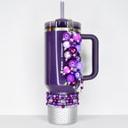 A purple Stanley + Starbucks tumbler with a handle, straw, and decorative beaded charm. The charm features various shades of purple beads. The bottom of the tumbler has a silver honeycomb design, and the base is adorned with beaded accents.