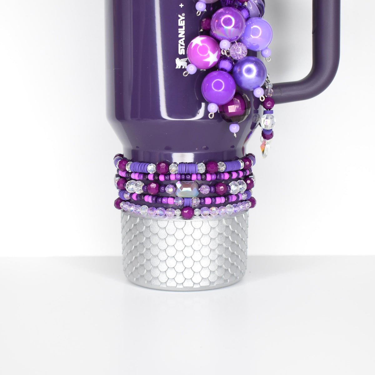 A purple Stanley tumbler with a decorative beaded design. The tumbler features a silver textured base and is adorned with various shades of purple beads and embellishments. The handle is a dark purple color.