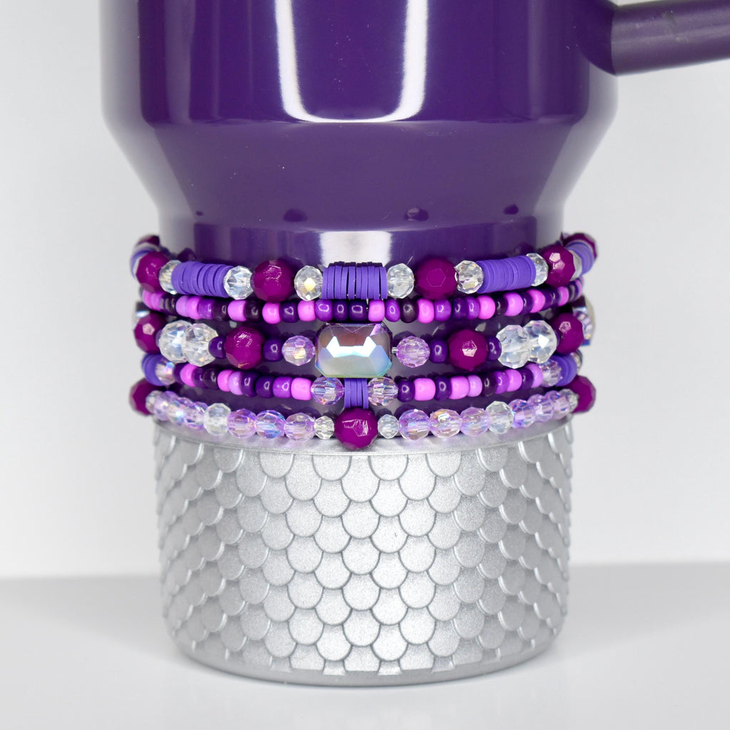 A plum Stanley with a silver, scale-patterned base. The tumbler is adorned with multiple layers of beaded bracelets in shades of purple, pink, and clear crystal. The bracelets feature various bead sizes and shapes, including a large, iridescent gem.