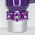 A plum Stanley with a silver, scale-patterned base. The tumbler is adorned with multiple layers of beaded bracelets in shades of purple, pink, and clear crystal. The bracelets feature various bead sizes and shapes, including a large, iridescent gem.