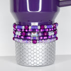A plum Stanley with a silver, scale-patterned base. The tumbler is adorned with multiple layers of beaded bracelets in shades of purple, pink, and clear crystal. The bracelets feature various bead sizes and shapes, including a large, iridescent gem.