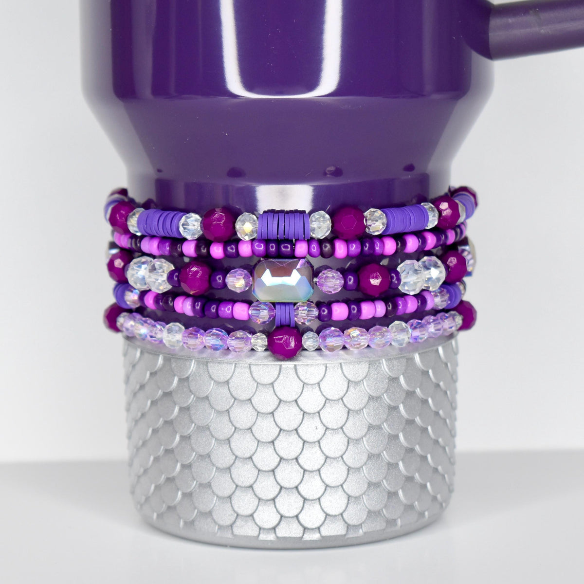 A plum Stanley with a silver, scale-patterned base. The tumbler is adorned with multiple layers of beaded bracelets in shades of purple, pink, and clear crystal. The bracelets feature various bead sizes and shapes, including a large, iridescent gem.