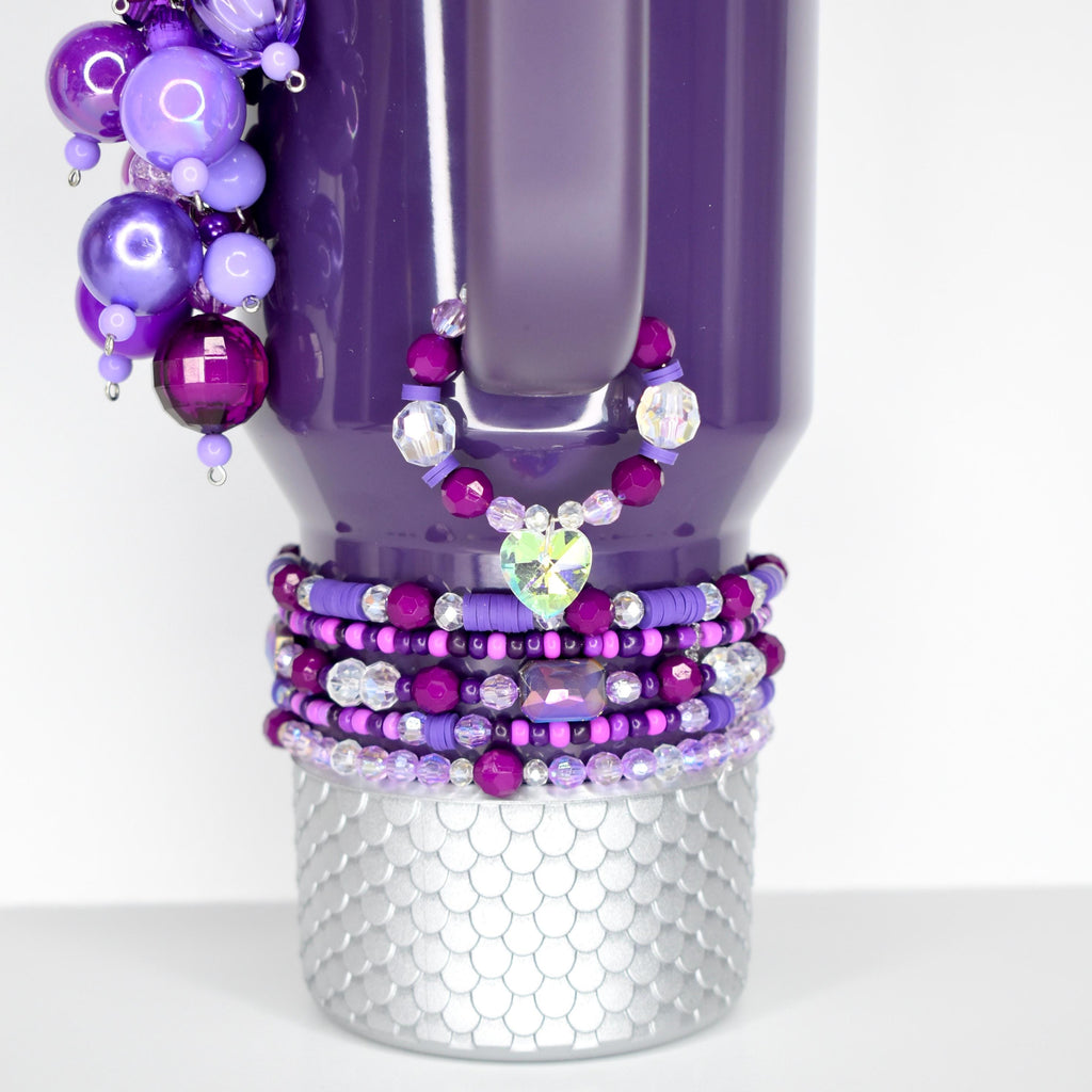 A purple tumbler with beaded embellishments. The tumbler features a silver, textured base and is adorned with various shades of purple beads and a heart-shaped charm. A cluster of purple beads hangs from the top.
