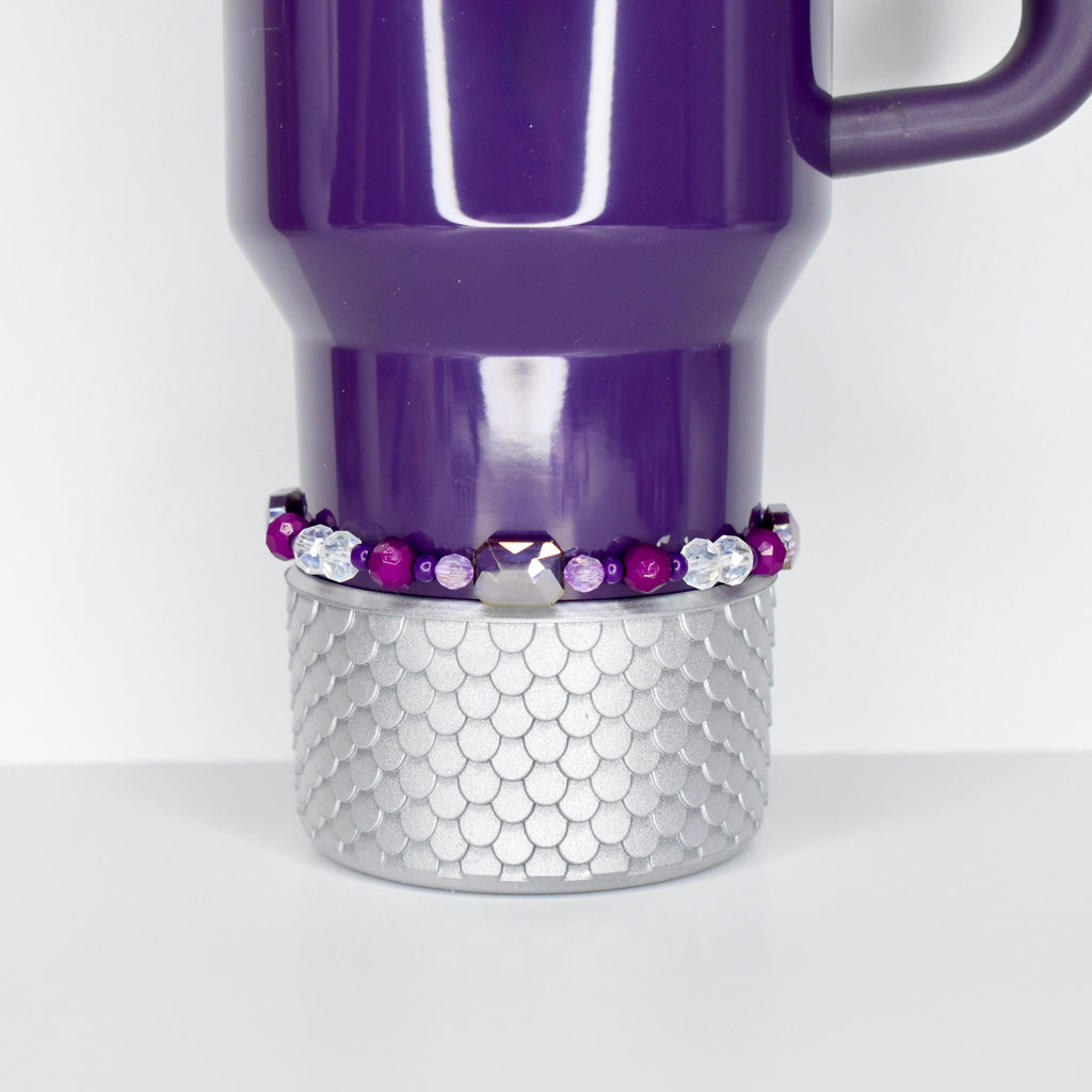 A purple Stanley with a silver, scale-patterned base. A beaded bracelet with purple and clear beads encircles the mug. The mug has a handle and a white interior.