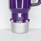 A purple Stanley with a silver, scale-patterned base. A beaded bracelet with purple and clear beads encircles the mug. The mug has a handle and a white interior.