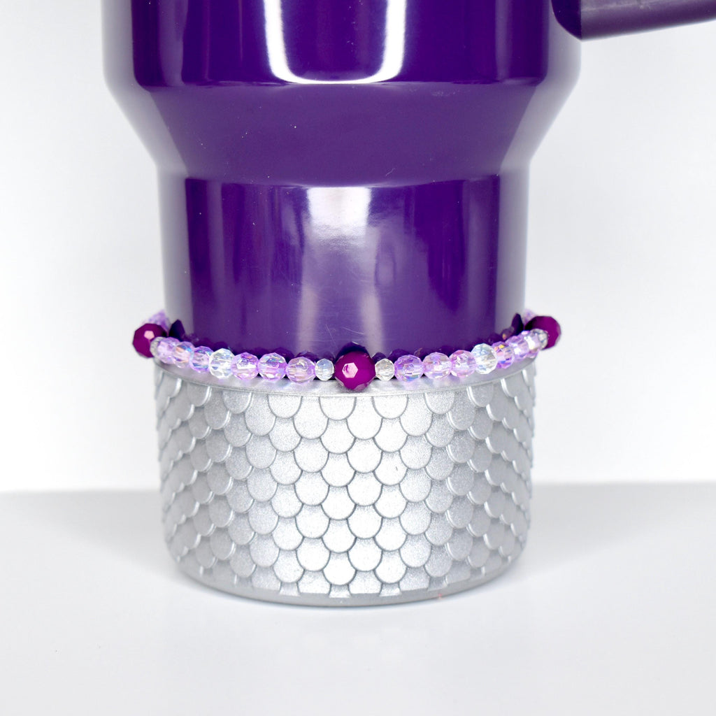A purple tumbler with a silver, scale-patterned base. A beaded bracelet with purple and clear beads encircles the tumbler. The tumbler has a handle and is set against a white background.