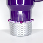 A purple tumbler with a silver, scale-patterned base. A beaded bracelet with purple and clear beads encircles the tumbler. The tumbler has a handle and is set against a white background.