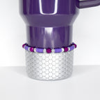 A purple Stanley tumbler with a silver base featuring a scale pattern. A beaded bracelet with purple and clear beads encircles the base. The tumbler has a handle and is set against a white background.
