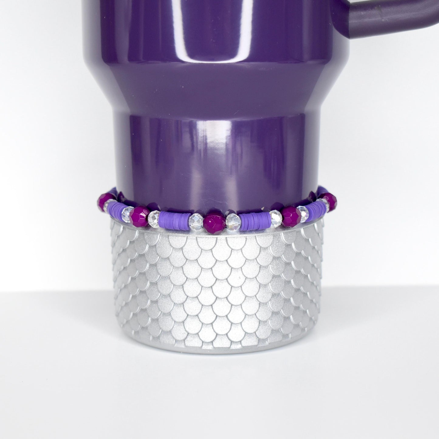 A purple Stanley tumbler with a silver base featuring a scale pattern. A beaded bracelet with purple and clear beads encircles the base. The tumbler has a handle and is set against a white background.