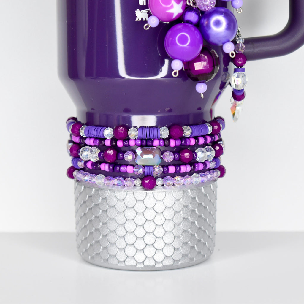 A purple mug with a silver, scale-patterned base. The mug is adorned with beaded bracelets in shades of purple and clear beads. A charm of purple beads and a crystal dangle from the handle.