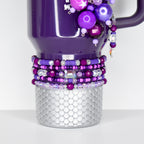 A purple mug with a silver, scale-patterned base. The mug is adorned with beaded bracelets in shades of purple and clear beads. A charm of purple beads and a crystal dangle from the handle.