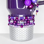 A purple tumbler with a silver, scale-like base. The tumbler is adorned with layers of purple and clear beads, and a large, iridescent gem. Additional purple beads and a charm hang from the handle.