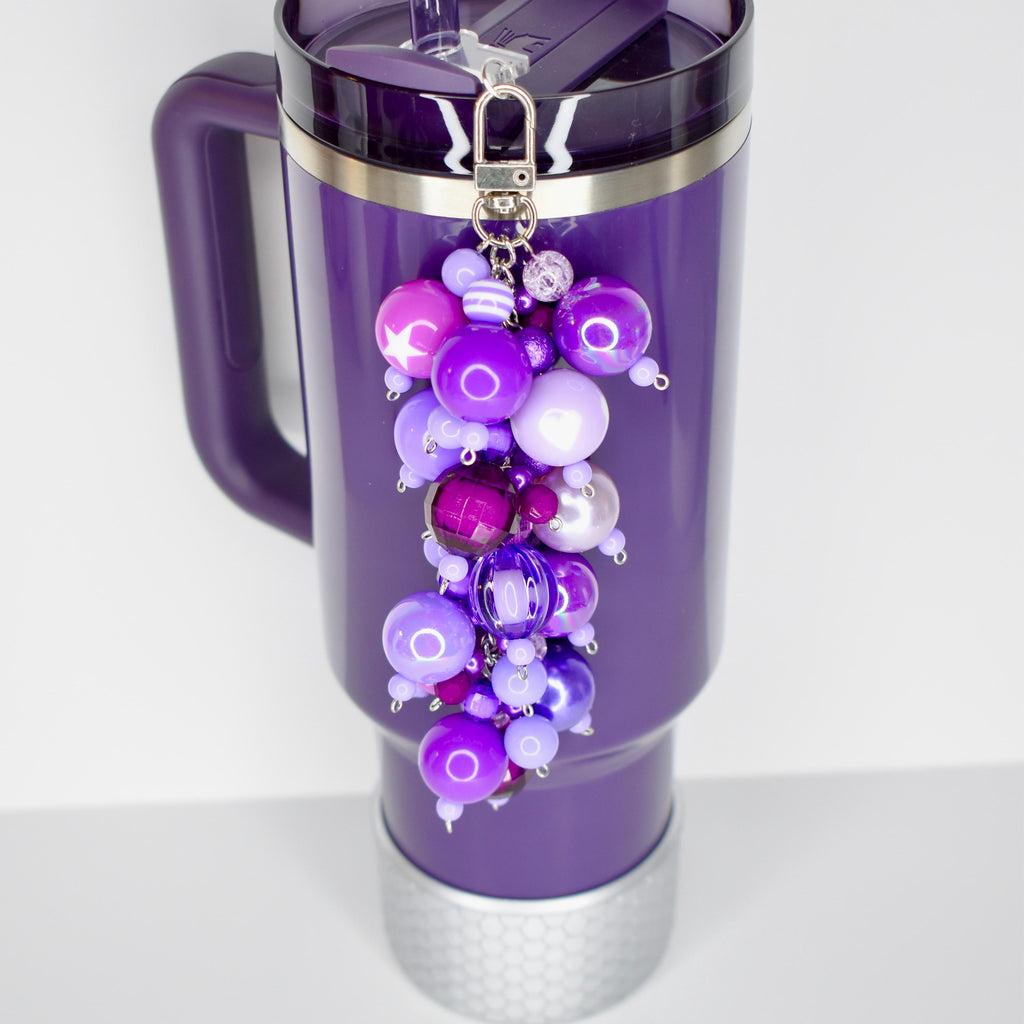A purple Stanley with a purple handle and a silver silicone boot at the base. A beaded straw charm in shades of purple, lavender, and silver hangs from the lid. The charm features various sized beads.
