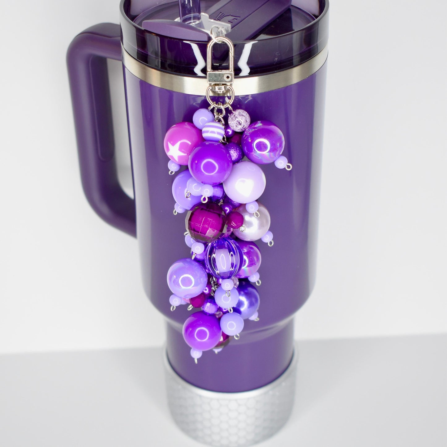 A purple Stanley with a purple handle and a silver silicone boot at the base. A beaded straw charm in shades of purple, lavender, and silver hangs from the lid. The charm features various sized beads.