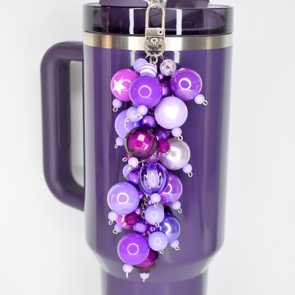 A plum Stanley with a silver clasp and a beaded straw charm. The charm features a cluster of purple beads in various shades and sizes, including a star-shaped bead. The mug has a handle and a dark purple lid.